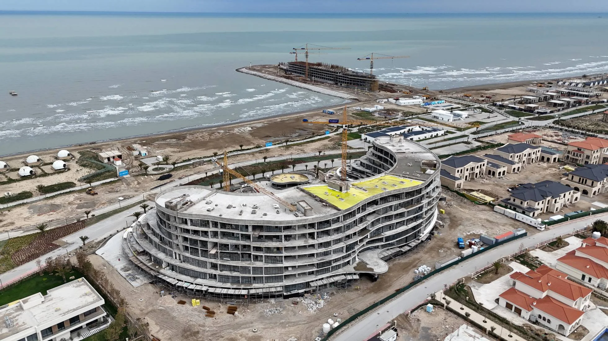 Skyline с высоты 18 апреля 2025 года: семь этажей каркаса подковой, два башенных крана, на кровле — жёлтая гидроизоляция. Слева у пляжа — белые купола кемпинга, в море на молу — стройка Caspian Dream Liner